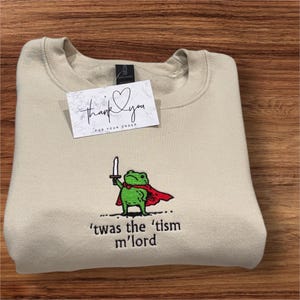May include: Beige sweatshirt with an embroidered green frog holding a sword and wearing a red cape. The text "'twas the 'tism m'lord" is below the frog. A small card with "thank you" and a heart is on top.