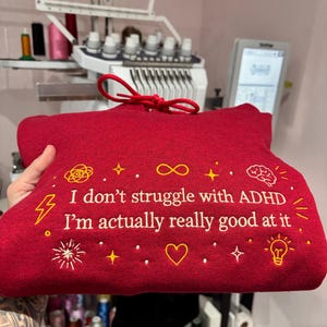 May include: A red hoodie with yellow embroidered text and symbols. The text reads "I don't struggle with ADHD I'm actually really good at it." The design includes a brain, lightning bolt, and other symbols.