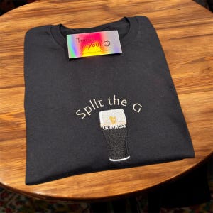 May include: Black t-shirt featuring an embroidered Guinness pint design. The design includes the text "Split the G" above the pint and the Guinness logo below. A small card with the text "Thank You!" is on top.