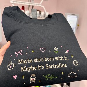 May include: Black sweatshirt with embroidered text and graphics. The text reads "Maybe she's born with it, Maybe it's Sertraline." The design includes a coffee cup, bow, heart, flower, pill, and other small icons.