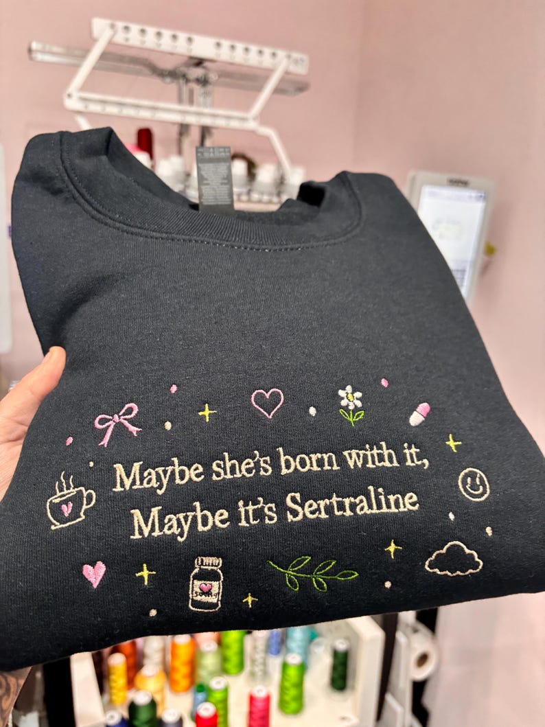 May include: Black sweatshirt with embroidered text and graphics. The text reads "Maybe she's born with it, Maybe it's Sertraline." The design includes a coffee cup, bow, heart, flower, pill, and other small icons.