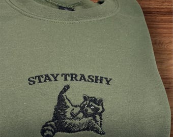 Puede incluir: Una sudadera verde oliva con la frase "STAY TRASHY" bordada en negro sobre una ilustración de mapache negro. El mapache está tumbado boca arriba. La sudadera está doblada sobre una superficie de madera.