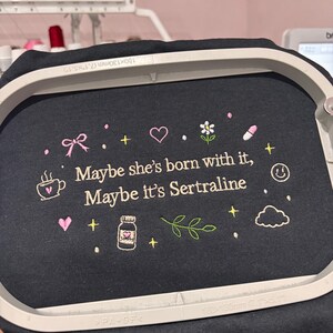 May include: Black fabric with embroidered text and graphics. The text reads "Maybe she's born with it, Maybe it's Sertraline." The design includes a bow, heart, flower, pill, coffee cup, bottle, cloud, and smiley face.