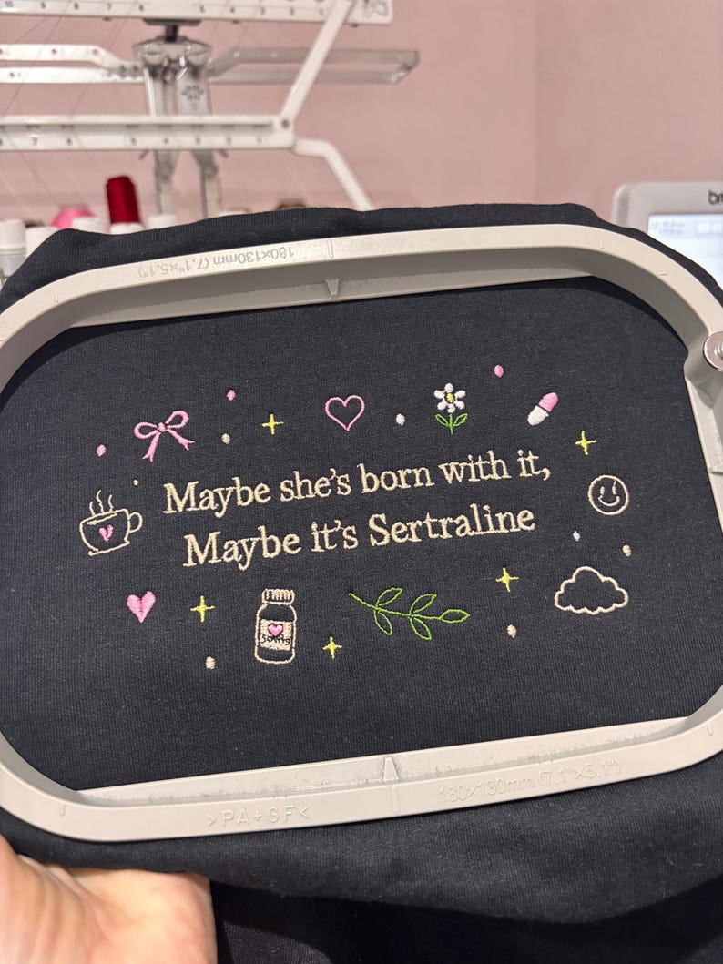 May include: Black fabric with embroidered text and graphics. The text reads "Maybe she's born with it, Maybe it's Sertraline." The design includes a bow, heart, flower, pill, coffee cup, bottle, cloud, and smiley face.