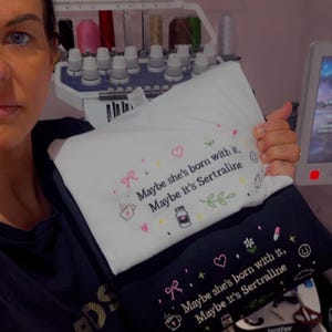 May include: Two embroidered pillowcases, one white and one black, with the phrase "Maybe she's born with it, Maybe it's Sertraline." The design includes a bow, heart, coffee cup, pill bottle, and leaves. The pillowcases are held in front of an embroidery machine.