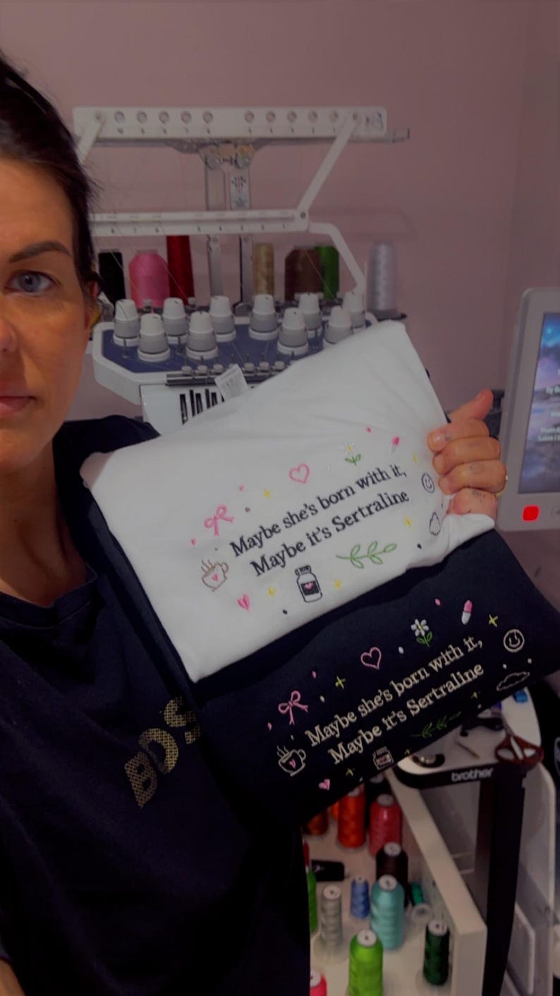 May include: Two embroidered pillowcases, one white and one black, with the phrase "Maybe she's born with it, Maybe it's Sertraline." The design includes a bow, heart, coffee cup, pill bottle, and leaves. The pillowcases are held in front of an embroidery machine.