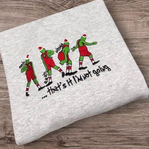 May include: A light grey sweatshirt featuring a graphic of four green figures wearing red overalls and Santa hats. The figures are in various poses with the text "ooh", "aaah", "whaaa", and "...that's it I'm not going" printed in black.