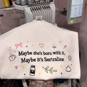 May include: White sweatshirt with embroidered text and graphics. The text reads "Maybe she's born with it, Maybe it's Sertraline." The design includes a coffee cup, heart, flower, pill, and other small icons.