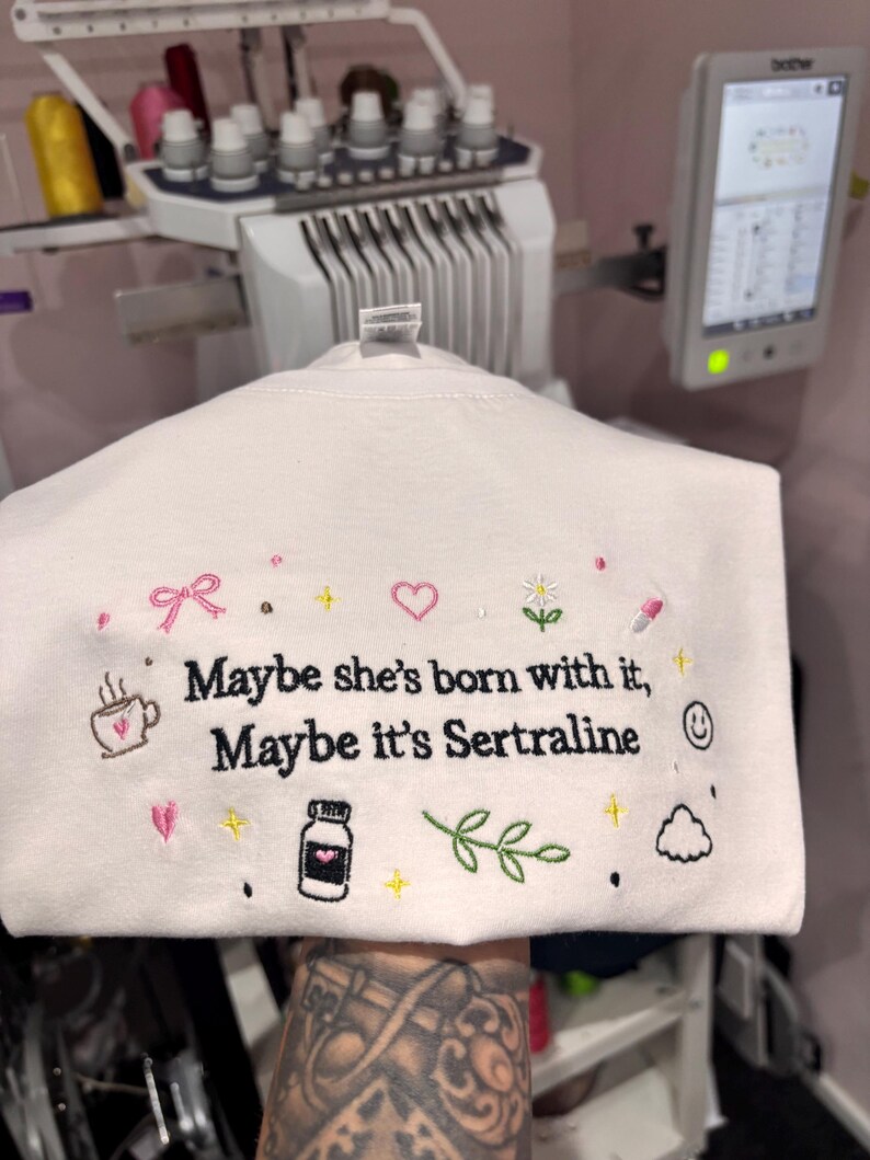 May include: White sweatshirt with embroidered text and graphics. The text reads "Maybe she's born with it, Maybe it's Sertraline." The design includes a coffee cup, heart, flower, pill, and other small icons.