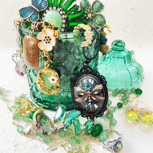 Puede incluir: Una colección de joyas verdes y doradas, que incluye collares, pendientes y anillos, exhibidas dentro y alrededor de un recipiente de vidrio verde. Las joyas presentan motivos de mariposas, flores y libélulas, con piedras preciosas y cristales verdes.