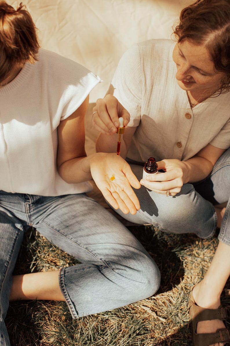 May include: Two people are seated outdoors, one holding a dropper with a reddish liquid, dispensing it into the other's palm. A small bottle is visible. Both are wearing casual clothing, including jeans and a white top. The scene is bathed in natural light.