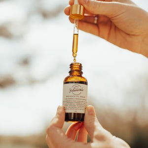 May include: A brown glass bottle of Radiance Oil Serum with a dropper held above it. The bottle has a white label with the text "Botanicals" and "Radiance Oil Serum". The dropper is gold and clear, filled with a golden liquid.