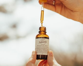 Puede incluir: Una botella de vidrio marrón de Radiance Oil Serum con un cuentagotas sostenido por encima. La botella tiene una etiqueta blanca con el texto "Botanicals" y "Radiance Oil Serum". El cuentagotas es dorado y transparente, lleno de líquido dorado.