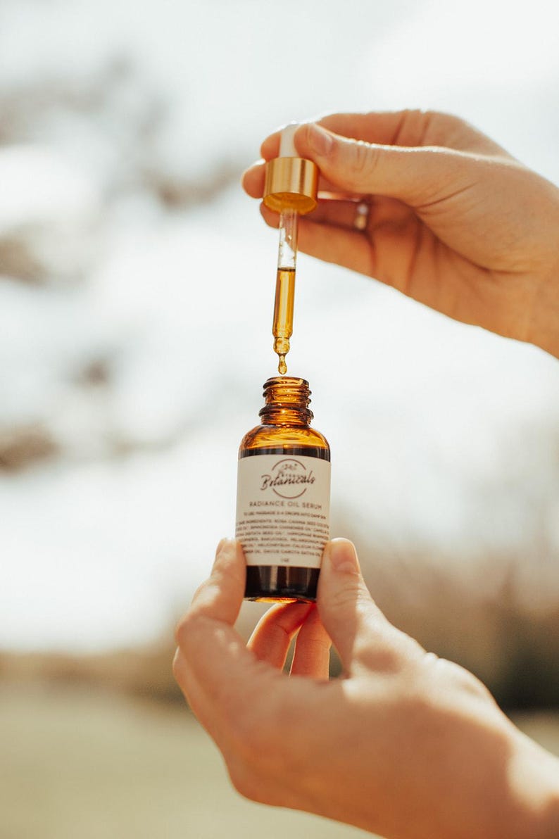 May include: A brown glass bottle of Radiance Oil Serum with a dropper held above it. The bottle has a white label with the text "Botanicals" and "Radiance Oil Serum". The dropper is gold and clear, filled with a golden liquid.