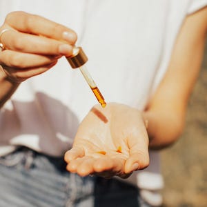May include: A close-up shot shows a dropper dispensing a golden-colored liquid onto a person's open palm. The dropper has a gold-colored top and a clear glass tube. The person is wearing a white shirt and blue jeans. The liquid appears to be an oil or serum.