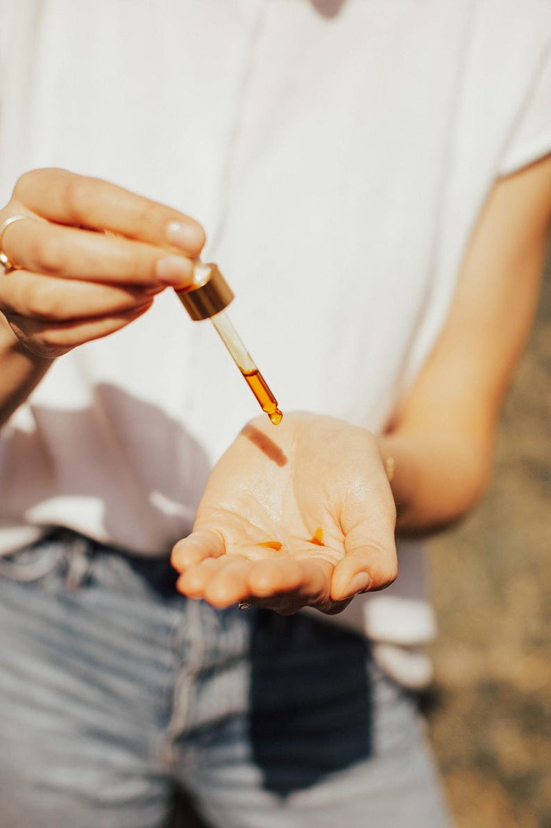 May include: A close-up shot shows a dropper dispensing a golden-colored liquid onto a person's open palm. The dropper has a gold-colored top and a clear glass tube. The person is wearing a white shirt and blue jeans. The liquid appears to be an oil or serum.