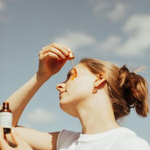 May include: A woman applying a golden-colored serum to her face with a dropper. She is wearing a white t-shirt and gold earrings. The background is a blue sky with clouds, suggesting an outdoor setting. A bottle of serum is held in her other hand.