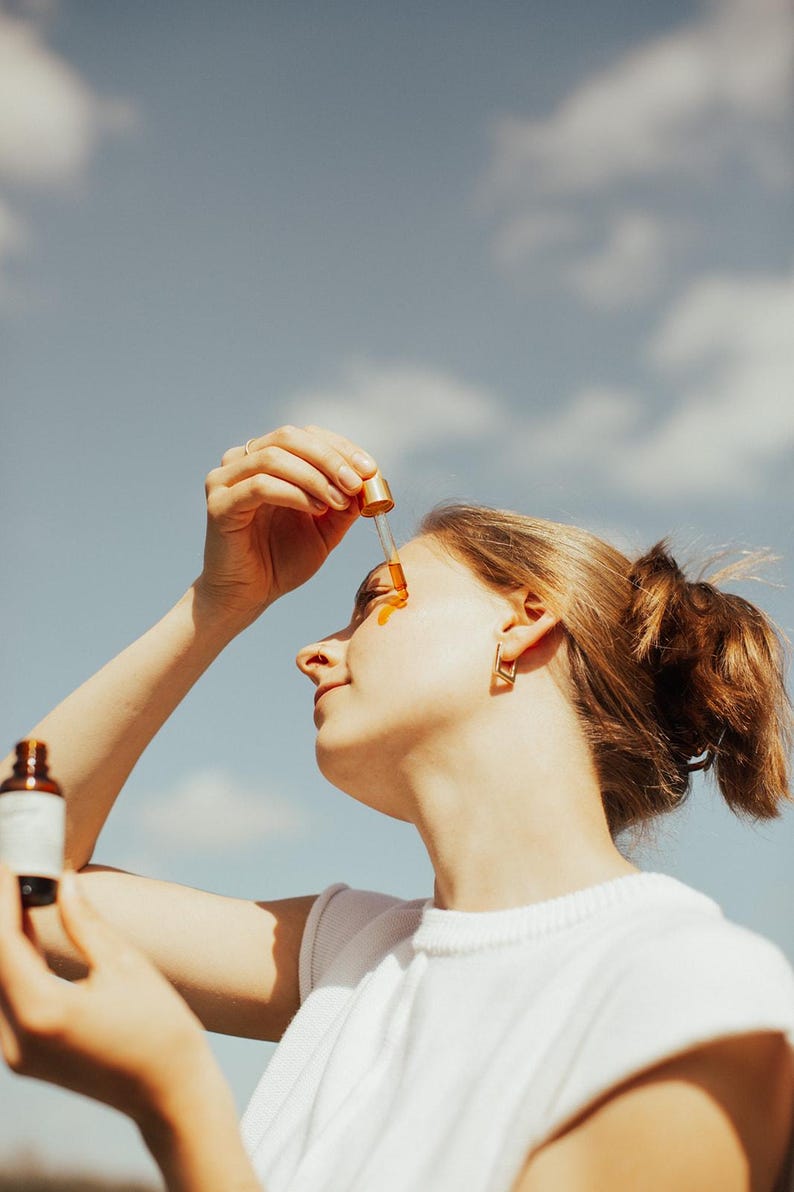 May include: A woman applying a golden-colored serum to her face with a dropper. She is wearing a white t-shirt and gold earrings. The background is a blue sky with clouds, suggesting an outdoor setting. A bottle of serum is held in her other hand.