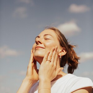 May include: A person with light skin wearing a white top, gold jewelry, and a nose ring, smiles with eyes closed, hands on face. The background is a blue sky with white clouds.