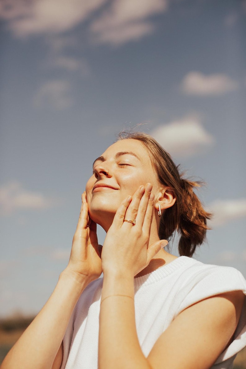 May include: A person with light skin wearing a white top, gold jewelry, and a nose ring, smiles with eyes closed, hands on face. The background is a blue sky with white clouds.
