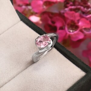 May include: A silver-toned ring featuring a single oval-cut light pink gemstone set in a prong setting. The band has a smooth, polished finish and a bypass design that curves around the central stone, presented in a black and beige jewelry box.