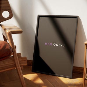 May include: A framed black poster with the text "MEN ONLY" in purple. The poster is leaning against a white wall, next to a wooden chair with a brown leather seat and a small wooden side table.