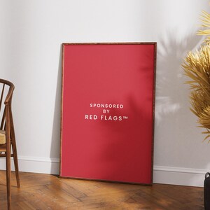 May include: A framed red poster with the text "SPONSORED BY RED FLAGS™" in white. The poster is leaning against a white wall on a wooden floor. A wooden chair and dried plants are also in the image.