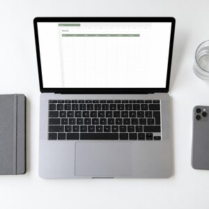 Könnte beinhalten: Ein silberner Laptop mit einer Tabellenkalkulation mit grünen Kopfzeilen, neben einem grauen Notizbuch, einem grauen Smartphone und einem klaren Glas Wasser. Der Bildschirm zeigt eine Tabelle mit den Spalten Name, Datum, Artikel, Status und Gesamt.