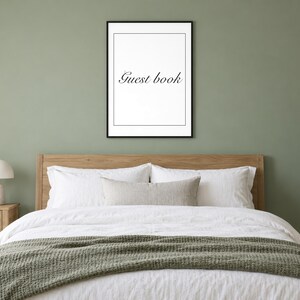 May include: A rectangular wall art print in a thin black frame featuring a white background with a thin black border and the text Guest book in a black serif font, centered and displayed on a solid sage green wall above a wooden bed frame.