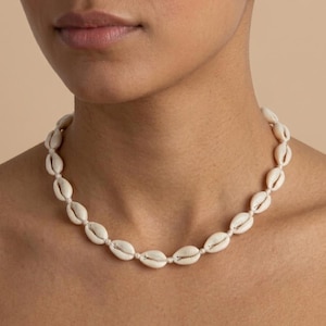 Puede incluir: Un collar de conchas de cauri de color crema. Las conchas están ensartadas con un fino cordón blanquecino. El collar se lleva alrededor del cuello. El fondo es de un color neutro sólido.