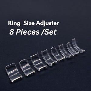 May include: A set of eight clear plastic ring size adjusters. The text "Ring Size Adjuster 8 Pieces / Set" is displayed above the adjusters. The adjusters are arranged in a row on a dark background.