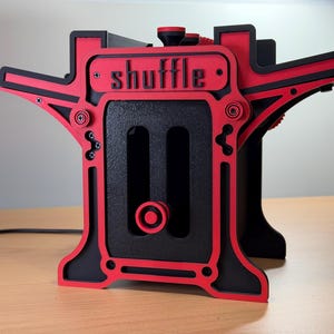 May include: A mechanical card shuffler featuring a black and red layered frame with the word shuffle embossed on the front. The device includes a central vertical slot with a red knob and exposed screws, constructed with a structured, angular design.