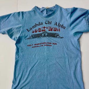 Può includere: T-shirt azzurra con la scritta "Lambda Chi Alpha ROCK 'N RUN" in rosso e nero. Presenta una grafica di un edificio e la scritta "HALF MARATHON/FUN RUN CORVALLIS, OREGON". La maglietta è vintage e ha un girocollo.
