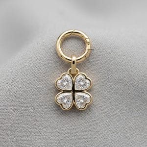 May include: A gold-toned charm featuring a four-leaf clover design composed of four heart-shaped clear stones. The charm is attached to a circular jump ring and includes the small text velousjewel printed near the top connector.