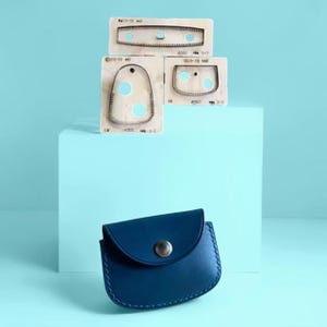 Könnte beinhalten: Ein marineblaues Leder-Münzportemonnaie mit silbernem Druckknopfverschluss liegt vor drei Stanzformen. Die Schablonen sind hell und haben verschiedene Formen mit kreisförmigen Aussparungen.
