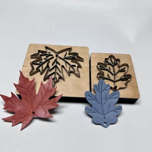 May include: Two wooden leaf-shaped die cut blocks with a red and a blue leaf-shaped cutout. The die cut blocks are made of light brown wood. The red leaf is a maple leaf shape, and the blue leaf is an oak leaf shape.