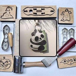 May include: A handmade panda bear keychain with a white and black design, a green backing, and a silver keyring. The image also shows various leather crafting tools and wooden stamps with Halloween-themed shapes.