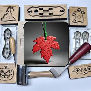 May include: A collection of leather crafting tools and supplies, including a red maple leaf pendant, wooden stamps with Halloween-themed designs, and various metal tools. The tools are arranged on a white surface.