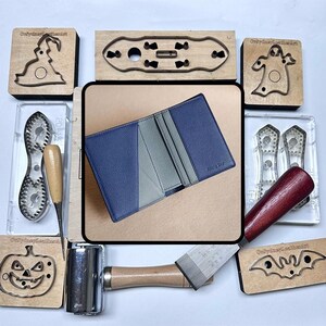 May include: A blue and grey leather wallet is surrounded by leatherworking tools and wooden stamps with Halloween-themed cutouts. The wallet is open, showing interior pockets. Other tools include a mallet and a chisel.