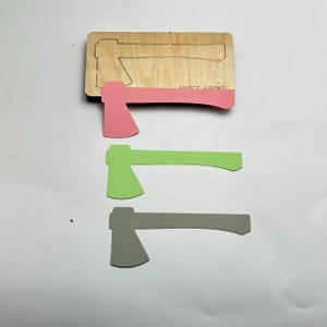 Puede incluir: Una matriz de corte de madera con formas de hacha, junto con tres recortes de hacha individuales en rosa, verde y gris. La matriz de corte tiene el texto "WUOCT 690MD".