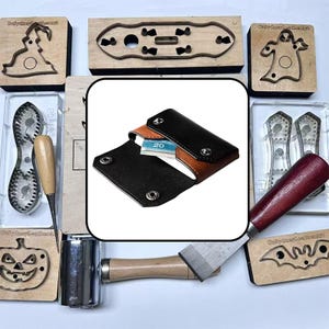 May include: A leather wallet with a black exterior and brown interior, featuring silver metal snap buttons and a folded twenty-dollar bill inside. The wallet is surrounded by various metal cutting dies and leatherworking tools on a white surface.