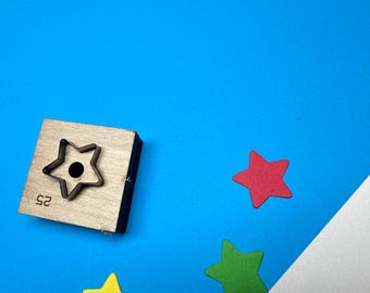 May include: A wooden star-shaped die-cut tool with a black star cutout and the number 25. Scattered around are three colorful star shapes in red, green, and yellow, against a blue background.