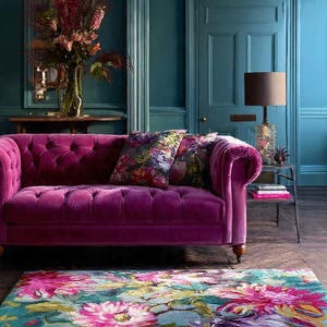 May include: A magenta velvet sofa with button tufting is the focal point of a room with teal walls. Floral throw pillows and a matching rug with pink and purple flowers add color. A table lamp and vase of flowers complete the scene.