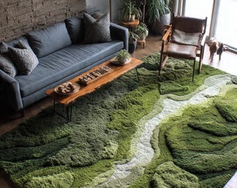 May include: A textured green and white rug with a landscape design. The rug's design uses varying shades of green and white to create a three-dimensional effect. The rug is in a living room with a grey sofa and wooden coffee table.