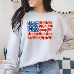 May include: A light gray sweatshirt featuring a watercolor design of the American flag. The flag's stars are represented by white dots on a blue background, and the stripes are replaced with red and pink flowers. The sweatshirt is made of a soft material.