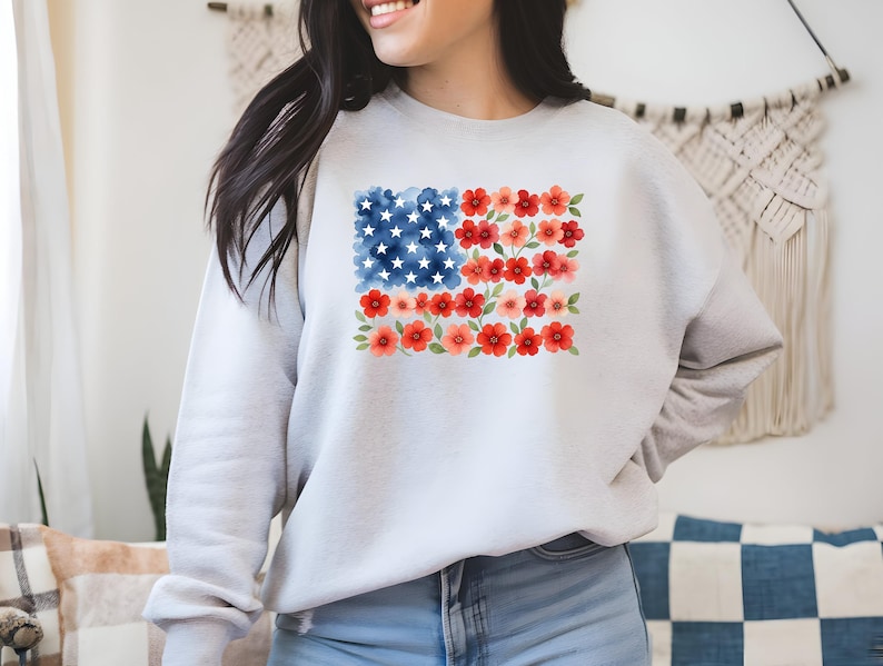 May include: A light gray sweatshirt featuring a watercolor design of the American flag. The flag's stars are represented by white dots on a blue background, and the stripes are replaced with red and pink flowers. The sweatshirt is made of a soft material.