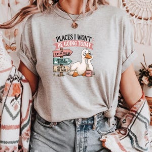 May include: A heather gray t-shirt featuring a graphic of a tired white duck holding a pink mug labeled NOPE. The text reads PLACES I WON'T BE GOING TODAY above three directional signs pointing to THE EXTRA MILE, ABOVE & BEYOND, and OUTTA MY WAY.