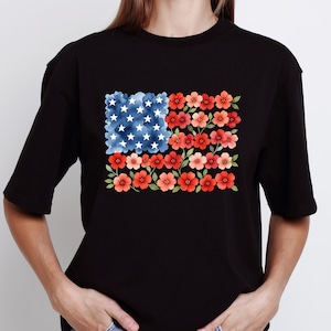 May include: Black t-shirt featuring a floral American flag design. The flag is composed of blue watercolor clouds with white stars and red and pink flowers. The shirt is a casual, everyday wear item.