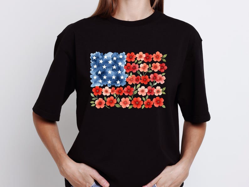 May include: Black t-shirt featuring a floral American flag design. The flag is composed of blue watercolor clouds with white stars and red and pink flowers. The shirt is a casual, everyday wear item.