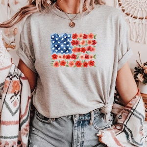 May include: A gray t-shirt featuring a watercolor design of the American flag. The flag design incorporates blue stars and red flowers. The shirt is paired with blue jeans and a patterned shawl.